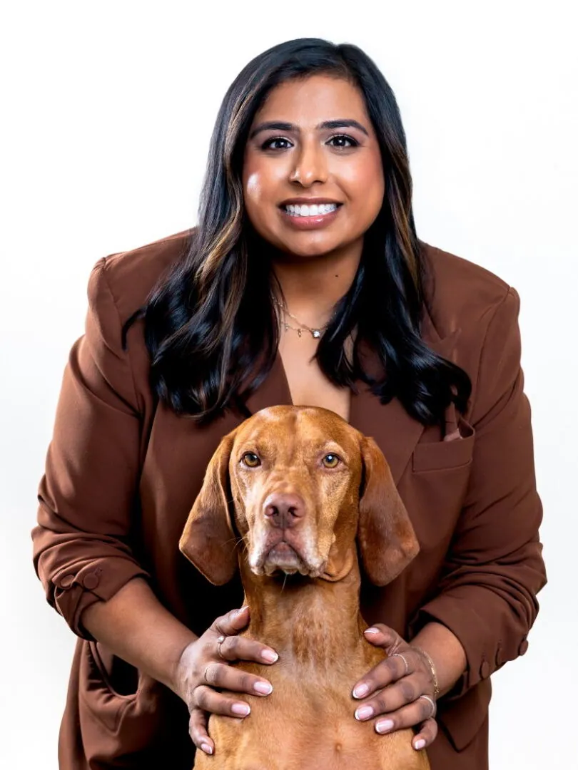 Dr. Anisha Jacob smiling behind Ollie, her vizsla, with her hands resting on the dog