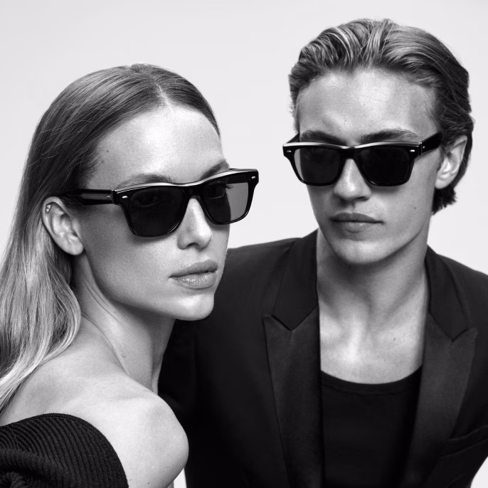 Black-and-white photo of a woman and man wearing Oliver Peoples black sunglasses