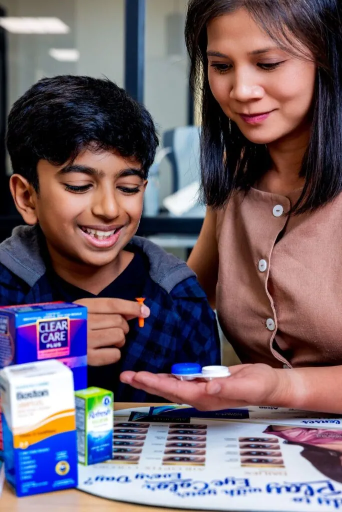 A Village Vision Center optometrist shows a smiling boy his ortho-k lens case and insertion tool, with lens-care products on the table