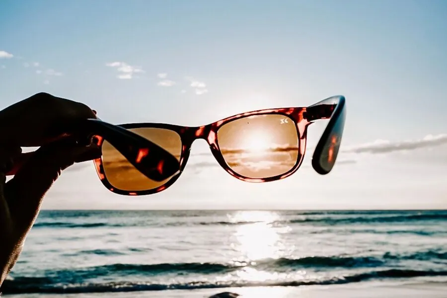 Hand holding up tortoiseshell sunglasses against the sun, with sunlight glaring off the ocean behind them