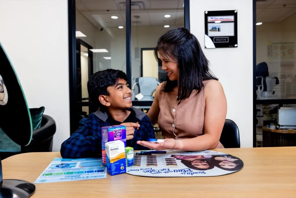 A young patient and a Village Vision Center team member smile at each other over contact lens solutions, a lens case and an Ortho-K care guide